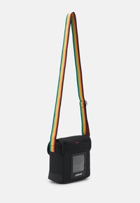 Sac bandoulière en tissu noir avec une poche transparente et une sangle rayée multicolore. Il présente un rabat supérieur et des détails en rouge.