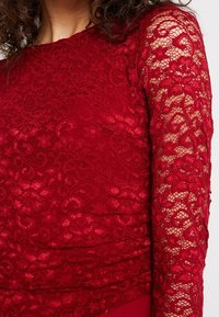 Top in pizzo rosso con maniche lunghe e trasparenti decorate con motivi floreali intricati e un corpino testurizzato, rifinito con un orlo inferiore liscio.