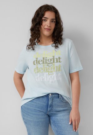 Personne aux cheveux bouclés portant un T-shirt bleu clair avec l'inscription « delight » et un jean en denim bleu, debout devant un fond gris clair uni.