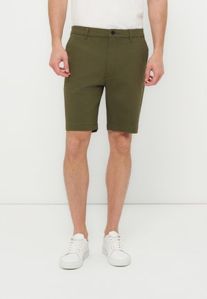 Egy férfi, aki olivazöld, térdig érő nadrágot, fehér alacsony szárú tornacipőt és fehér pólót visel, egy fehér háttér előtt áll.