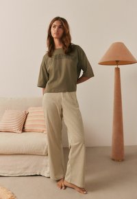 Olivgrünes, kurz geschnittenes T-Shirt mit dem schwarzen Schriftzug "Copenhagen", kombiniert mit beigefarbenen und braunen, karrierten Hosen mit weitem Bein. Hintergrund in neutralen Tönen.