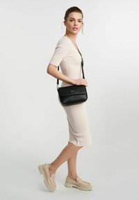 Czarna skórzana torebka na ramię z klapką i srebrnymi elementami. Umieszczona na modelce w dopasowanej, żebrowanej beżowej sukience i kremowych butach.