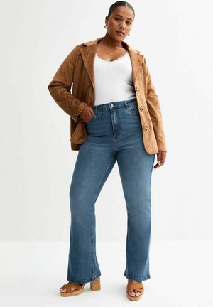 Giacca in suede marrone, canotta bianca, jeans a vita alta svasati blu e sandali con zeppa beige. Texture lisce con dettagli di bottoni e tasche.