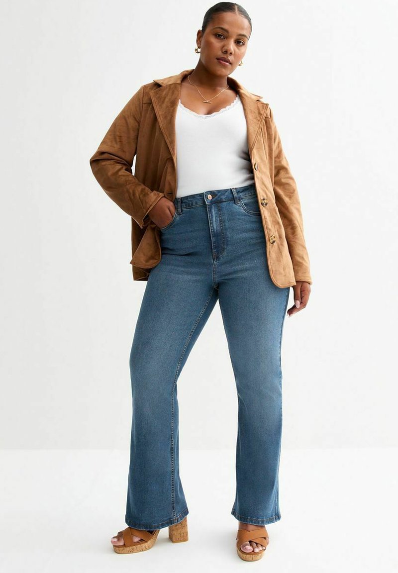 Brun mockaskinnjacka, vit linne, högmidjade vida blå jeans och beige platåsandaler. Släta texturer med knapp- och fickdetaljer.