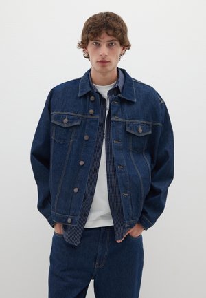 Giubbotto di jeans blu scuro con due tasche sul petto, bottoni in metallo e cuciture a contrasto; indossato sopra una camicia a scacchi e una T-shirt bianca.