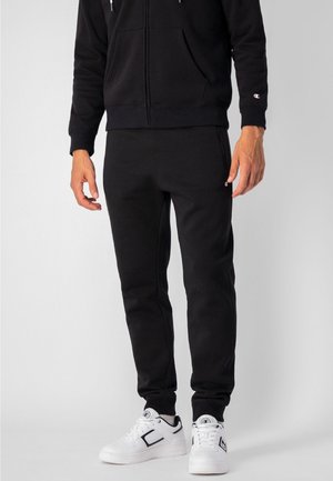 Sudadera negra con bolsillos frontales y cordón, combinada con pantalones de chándal negros, llevados con zapatillas blancas con acentos en negro.