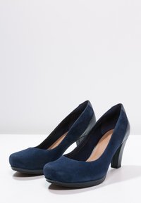 Námořnické semišové lodičky na vysokém podpatku s kulatou špičkou a kontrastním černým lakovaným podpatkem; hladký povrch a minimalistický design.