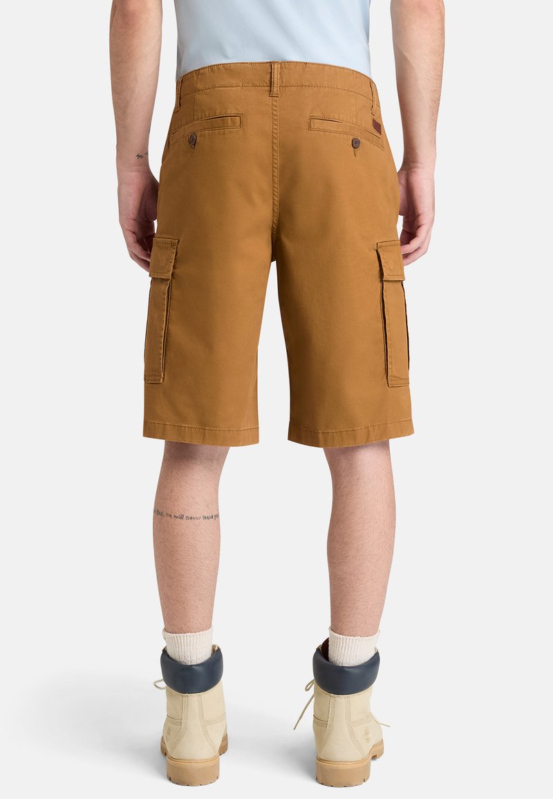 Man die bruine cargoshorts draagt, crèmekleurige laarzen met donkere kragen, witte sokken en een lichtblauw shirt, gezien van achteren.