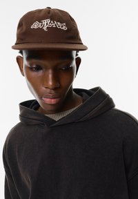 Brun keps med vit brodering "OUTLAWS", bärs med en mörkbrun hoodie med en texturerad tyg och en ribbad krage.