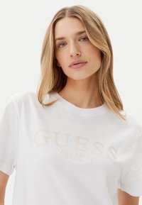 T-shirt bianco in cotone con la scritta "GUESS" in morbido ricamo rosa chiaro e "Los Angeles" sottostante, con collo girocollo standard e maniche corte.