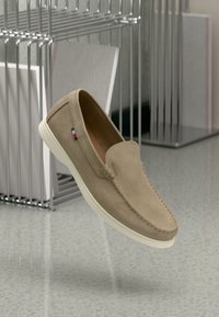 Tan suede slip-on shoes with white rubber sole, featuring subtle stitching and a branded accent on the side. Lightweight and contemporary design.