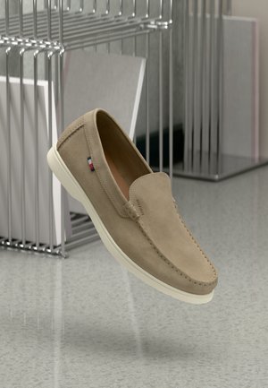 Beżowe zamszowe buty typu slip-on z białą gumową podeszwą, charakteryzujące się subtelnym przeszyciem i markowym akcentem z boku. Lekkie i nowoczesne w designie.