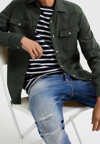 Camisa de botones verde oliva con dos bolsillos en el pecho, superpuesta a un suéter a rayas navy y blanco. Llevada con jeans rotos azul claro.