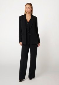 Completo nero su misura composto da un blazer con rever a punta e un gilet coordinato, abbinato a pantaloni a gamba larga e décolleté in vernice.