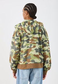 Sudadera con diseño de camuflaje en verde, marrón y gris, que cuenta con capucha con cordón, puños acanalados y un corte holgado. Textura de tela suave.