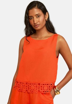 Frau mit langen dunklen Haaren trägt ärmelloses, leuchtend oranges Top mit floraler Spitzenverzierung und passende Hose, ergänzt durch goldene Armbänder.