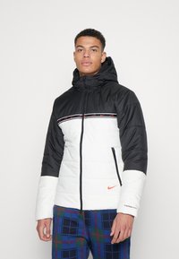 Chaqueta tipo puffer de Nike con capucha negra, cuerpo blanco y detalles rojos. Cuenta con cierre de cremallera, una franja horizontal con el logotipo y puños elásticos.