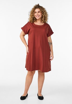 Usmievavá žena s kučeravými vlasmi v voľných šatách farby hrdzavej a čiernych balerínach, stojaca s jednou rukou v vrecku na bielom pozadí.