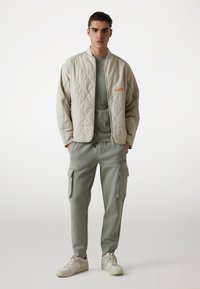 Beżowa pikowana kurtka z pomarańczowym logo, w zestawieniu z jasnooliwkowymi spodniami cargo z bocznymi kieszeniami oraz białymi trampkami. Minimalistyczny design.