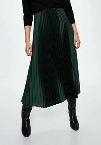 Green pleated skirt, made of lightweight fabric, features vertical pleats, a flowy design, and an elastic waistband, paired with black knee-high boots.