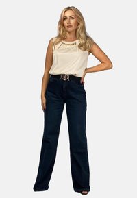 Kremowy top bez rękawów z łańcuszkiem, zestawiony z ciemnoniebieskimi spodniami o szerokich nogawkach z wysokim stanem oraz czarnym paskiem z metalową klamrą.