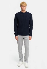 Pull en maille torsadée marine avec poignets et ourlet côtelés, associé à un pantalon de survêtement gris clair fuselé et des baskets blanches sur un fond uni.