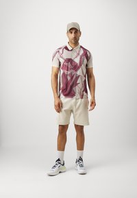 Beige spalvų trumpų rankovių polo marškiniai su geometriniu raštu bordo spalvos. Suderinti su beigių spalvų šortais ir baltais sportbačiais su juodais akcentais. Kepurė užbaigia aprangą.