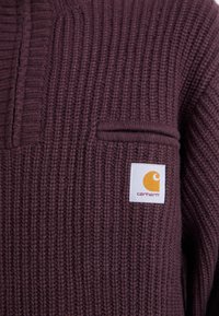 Temno purpurna pletena majica z rebrasto teksturo, z majhnim žepom na prsih in oznako Carhartt v rumeni in beli barvi.