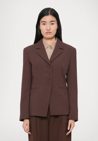 Hnedé na mieru ušité sako s výstrihom do vrúbka, predné zapínanie na päť gombíkov a dve bočné vrecká, doplnené béžovou košeľou s golierom pod ním.