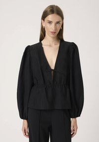 Musträp must blouse sügava V-kaelusega, pikkade puffvarrukatega ja kinnitatud taljega. Tekstureeritud kangas vertikaalsete triipudega ja lipsutähtsusega kaeluses.