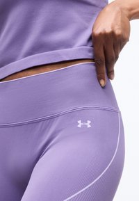 Μέση και γοφοί ατόμου που φοράει μωβ κολάν Under Armour και αντίστοιχο τοπ, με το χέρι να τραβάει ελαφρώς τη ζώνη της μέσης μακριά από το σώμα.