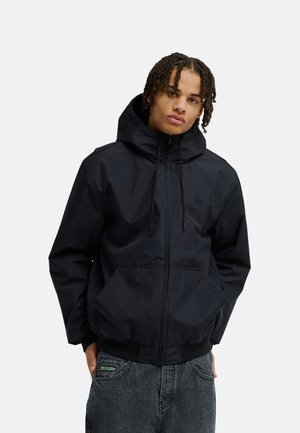 Junge Person mit geflochtenem Haar, trägt eine schwarze Kapuzenjacke und dunkle Jeans, die Hände teilweise in den Taschen.