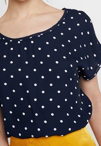 Námořnická polka dot blůza s krátkými rukávy, kulatým výstřihem a bílým tečkovaným vzorem. Tento kousek má měkkou, splývavou tkaninu a volný střih.