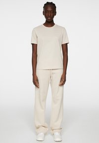 Beige bomullströja med korta ärmar, rund halslinning och en subtil logotyp; matchad med beiga sweatpants och vita sneakers.