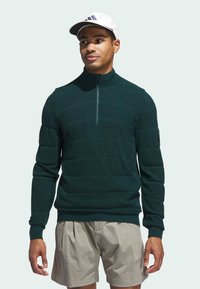 Mørkegrønn jakke med glidelås og horisontale striper, laget av strukturert stoff, kombinert med lyse beige shorts. Klassisk atletisk design.