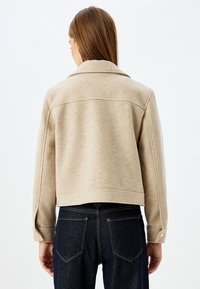 Giacca in misto lana beige con colletto classico, spalle scese e silhouette cropped. Presenta polsini con bottoni e dettagli sulle cuciture del petto.