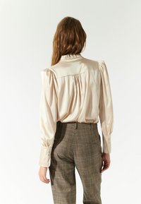 Blusa de satén crema con mangas abullonadas, cuello con volantes y detalle fruncido en la parte trasera, combinada con pantalones marrones a cuadros.