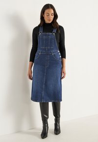 Denim overall klänning, mörkblå, midi längd, framfickor, justerbara axelremmar, silver-tonad hårdvara, ihopkopplad med svarta knähöga stövlar.