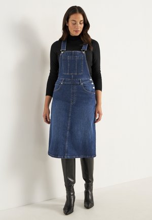 Denim overall kjole, mørkeblå, midi længde, frontlommer, justerbare skulderstropper, sølvfarvet hardware, kombineret med sorte knæhøje støvler.