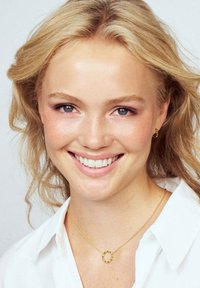 Närbild av en person med ljust blont, vågigt hår. De bär ett guldpläterat cirkulärt hänge och små guldörhängen, mot en ljus bakgrund.