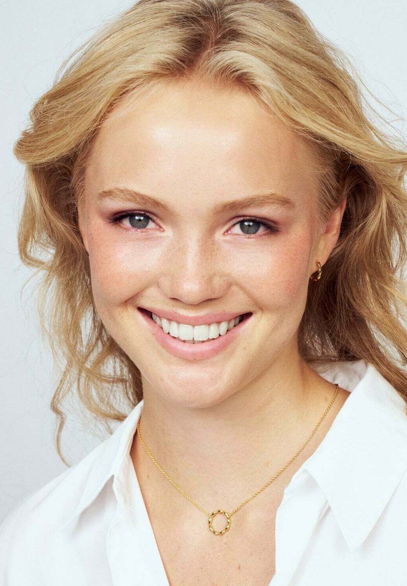 Närbild av en person med ljust blont, vågigt hår. De bär ett guldpläterat cirkulärt hänge och små guldörhängen, mot en ljus bakgrund.