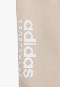 Béžová látka s hladkou texturou, na které je velké bílé logo "adidas" a text "SPORTSWEAR" vertikálně zarovnaný po straně.