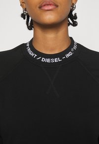 Svart sweatshirt med ribbad krage och vit "DIESEL"-logotyp. Den har en slät textur och en rundad halslinning med en korsstygnsdetalj.