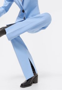 Person wearing light blue suit pants with side slits and black heeled boots, posing with one leg bent and lifted.