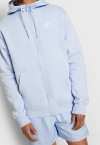 Ljusblå zip-up huvtröja i mjukt tyg, med en känguruficka, dragsko i huvan och vit Nike-logotyp på övre vänster.