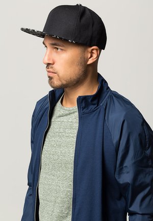 Black cap with patterned brim, navy zip-up jacket, and light gray shirt. Features a modern design with a smooth texture and fitted shape.