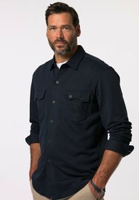 Camicia bottonata a maniche lunghe color navy, in tessuto testurizzato, con due tasche sul petto dotate di pattina e bottoni, polsini arrotolati, indossata sopra una t-shirt bianca.