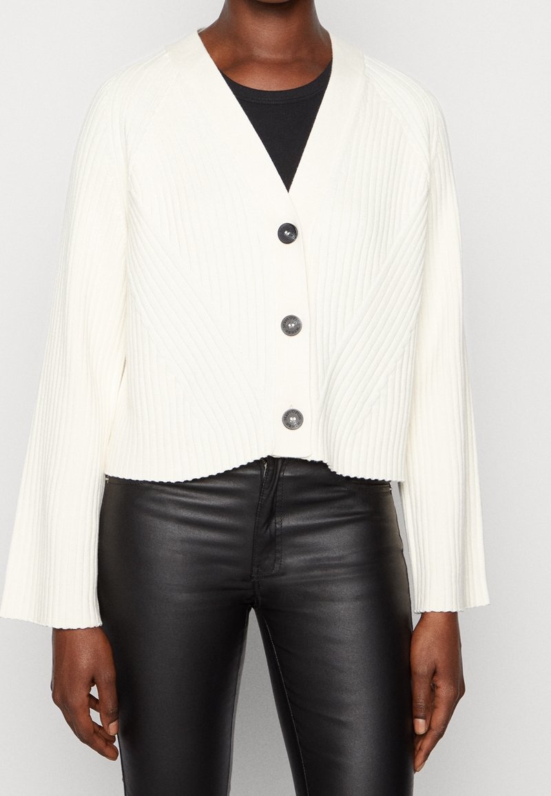 Avklippt vit ribbad cardigan med V-ringning, tre mörka knappar och vida ärmar, i kombination med svarta läderbyxor. Texturerad tygdetalj.