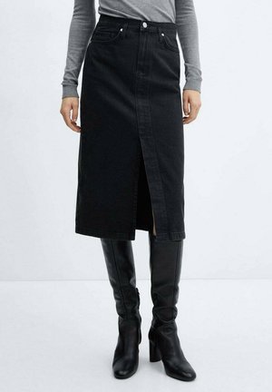 Kvinde iført en sort midi denimnederdel med frontslids, grå langærmet top og sorte knæhøje støvler med hæl foran en ensfarvet baggrund.