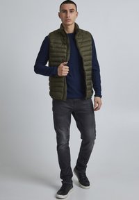 Zeleni puffer brez rokavov nad temno modro srajco z dolgimi rokavi, v kombinaciji s sivimi kavbojkami in črnimi supergami. Gladka tekstura, zadrga in stranski žepi.
